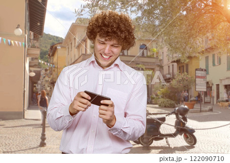 Emotional young man with curly hair playing a game using his smartphone. Urban city street in the background. 122090710