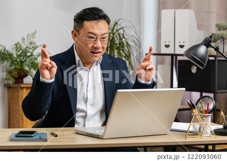 Asian businessman crossed fingers looking at laptop screen asking for good luck news win victory Asian businessman crossed fingers looking at laptop screen asking for good luck news win victory 122093060