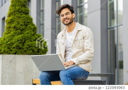 Indian man freelancer working on laptop computer sends messages reading email outdoors on bench Indian man freelancer working on laptop computer sends messages reading email outdoors on bench 122093063