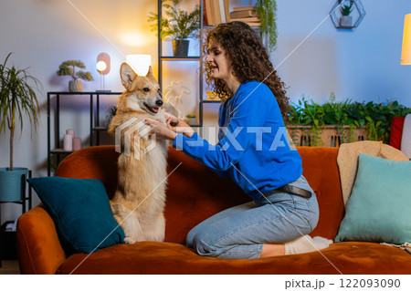 Happy smiling young woman playing with cute funny adorable corgi dog on sofa in room at home 122093090