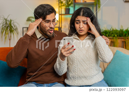 Disappointed multiethnic couple man woman reading together bad news on smartphone at home on sofa 122093097