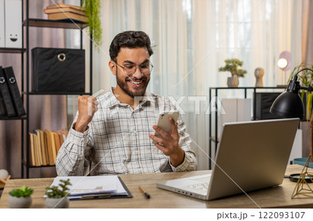 Happy Indian businessman using smartphone celebrating success victory winning lottery casino jackpot Happy Indian businessman using smartphone celebrating success victory winning lottery casino jackpot 122093107
