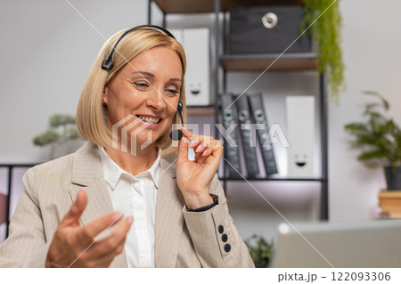 Confident mature businesswoman in headset talking on video call using laptop at home office desk 122093306