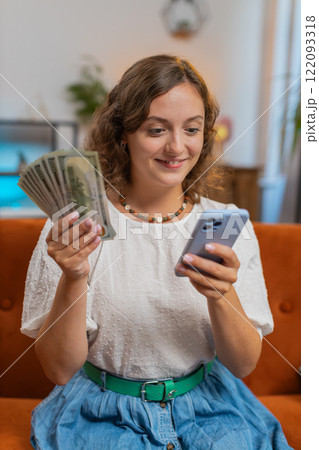 Successful young woman holding smartphone and dollar bill money fan sitting on sofa couch at home Successful young woman holding smartphone and dollar bill money fan sitting on sofa couch at home 122093318