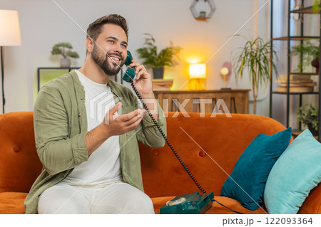 Smiling young man making wired telephone call conversation with friends sitting on sofa at home 122093364