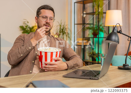 Happy young businessman taking break from work eating popcorn and watching movie on laptop at home Happy young businessman taking break from work eating popcorn and watching movie on laptop at home 122093375