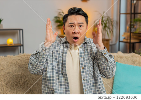 Upset Chinese man making face disappointed in result, bad news watching TV on sofa couch at home Upset Chinese man making face disappointed in result, bad news watching TV on sofa couch at home 122093630