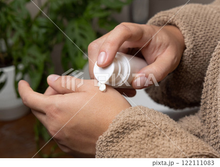 Woman in brown bathrobe applying cream on hand and holding a cosmetic bottle, closeup in bathroom Woman in brown bathrobe applying cream on hand and holding a cosmetic bottle, closeup in bathroom 122111083