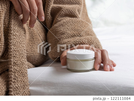 Woman in brown bathrobe puts white opened cream jar on white bedsheet closeup, mockup Woman in brown bathrobe puts white opened cream jar on white bedsheet closeup, mockup 122111084
