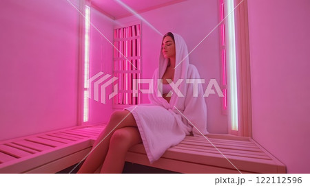 Young woman is sitting on a wooden bench, relaxing in an infrared sauna. 122112596