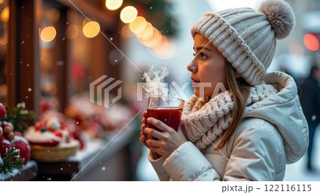 A girl, dressed in a white knitted hat and jacket, stands by a window display and drinks a warm beverage at a Christmas market 122116115