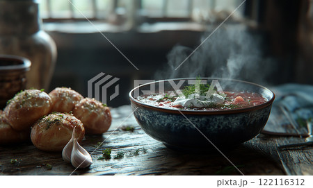 Russian Borscht bowl of soup with a dollop of sour cream on top sits on a wooden table. The bowl is filled with a hearty stew, and there are two rolls of bread nearby. 122116312