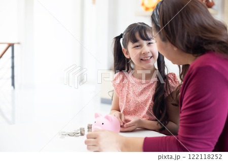 Smiling mother helping daughter putting money in piggy bank, Cute little girl saving money by adding a coin in piggy bank with mother at home. Smiling mother helping daughter putting money in piggy bank, Cute little girl saving money by adding a coin in piggy bank with mother at home. 122118252