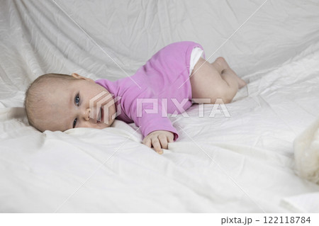 Cute and beautiful 4-month-old baby girl laying on bed and look at the camera. Infant in pink bodysuit on bed against white background. 122118784