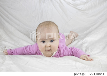 Cute and beautiful 4-month-old baby girl laying on bed and look at the camera. Infant in pink bodysuit on bed against white background. 122118841
