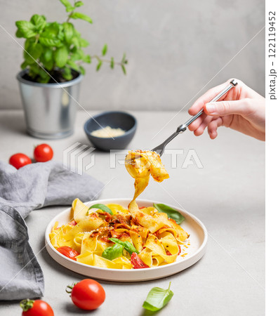 Hand holding fork with tagliatelle or pappardelle pasta with tomato sauce, basil and parmesan cheese Hand holding fork with tagliatelle or pappardelle pasta with tomato sauce, basil and parmesan cheese 122119452