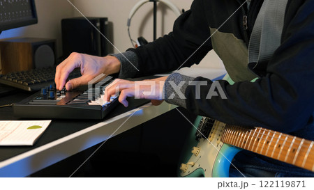 Musician sits at his desk, playing a MIDI keyboard with expertise while holding a mint electric guitar, fully immersed in songwriting and music production Musician sits at his desk, playing a MIDI keyboard with expertise while holding a mint electric guitar, fully immersed in songwriting and music production 122119871