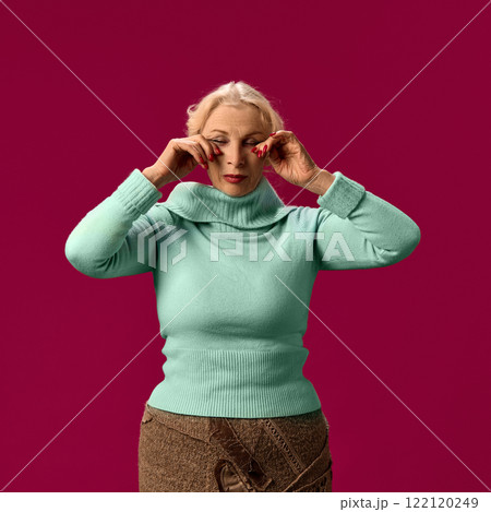 Senior woman in mint sweater swiping tears, suffering from seasonal allergy symptoms on on pink studio background 122120249