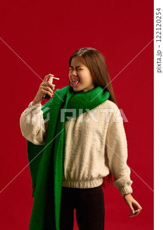 Teen girl in green scarf and beige sweater suffering from cold and flu, using throat spray to reduce soreness isolated on red background. Teen girl in green scarf and beige sweater suffering from cold and flu, using throat spray to reduce soreness isolated on red background. 122120254