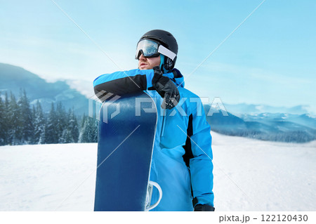 Snowboarder Of Man At Ski Resort On The Background Blue Sky, Hold Snowboard. Wearing Ski Glasses. Ski Goggles With The Reflection Of Snowed Mountains Snowboarder Of Man At Ski Resort On The Background Blue Sky, Hold Snowboard. Wearing Ski Glasses. Ski Goggles With The Reflection Of Snowed Mountains 122120430