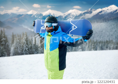 Portrait Of Man On The Background Of Mountains And Blue Sky, Holding Snowboard And Wearing Ski Glasses. Ski Goggles Of Snowboarder With The Reflection Of Snowed Mountains. Winter Sports 122120431