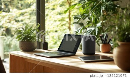 Laptop, tablet, and smart speaker on a wooden desk in a sunlit room with plants. 122121971