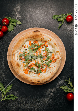Gourmet pizza with smoked salmon, arugula, capers, and melted cheese on a wooden plate Gourmet pizza with smoked salmon, arugula, capers, and melted cheese on a wooden plate 122122554