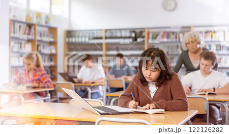 Schoolgirl studies on a laptop and writes notes in a copybook Schoolgirl studies on a laptop and writes notes in a copybook 122126282