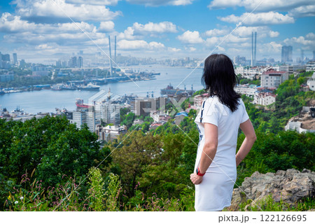 A 50-year-old Russian woman in a white dress stands in Nagorny Park, Vladivostok, admiring the view of Golden Horn Bay and the cityscape 122126695