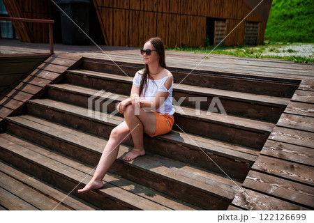 A barefoot Russian woman in her 40s sits on wooden stairs in summer, wearing orange shorts and a white blouse. She enjoys the sunny weather with a relaxed expression A barefoot Russian woman in her 40s sits on wooden stairs in summer, wearing orange shorts and a white blouse. She enjoys the sunny weather with a relaxed expression 122126699