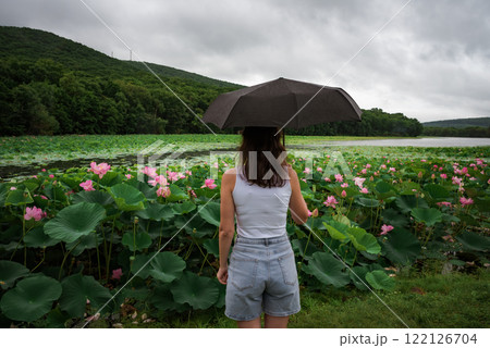 A young Russian woman with an umbrella stands alone by a lotus lake under a cloudy sky, symbolizing solitude, melancholy, and the mood of traveling in rainy weather A young Russian woman with an umbrella stands alone by a lotus lake under a cloudy sky, symbolizing solitude, melancholy, and the mood of traveling in rainy weather 122126704