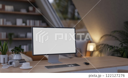 A close-up of a computer desk in a cozy, contemporary bedroom features a computer with a white screen mockup. 122127276