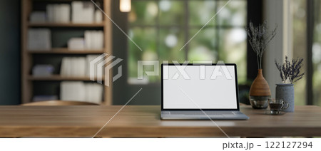 A laptop with a white screen mockup placed on a wooden table in a cozy contemporary room. 122127294
