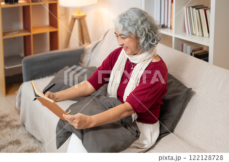 A happy Asian senior retired woman enjoys writing in her dairy on a sofa in the living room. A happy Asian senior retired woman enjoys writing in her dairy on a sofa in the living room. 122128728