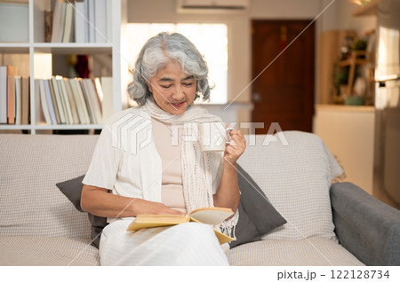 A well-being, happy Asian senior retired woman reads a book and enjoys a hot drink on a sofa. 122128734
