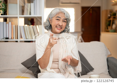 A well-being Asian senior woman, sits on a sofa, pointing with a gesture and smiling at the camera. 122128745