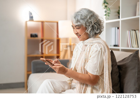 A happy Asian senior woman uses her smartphone to chat on social media while sitting on a sofa. A happy Asian senior woman uses her smartphone to chat on social media while sitting on a sofa. 122128753