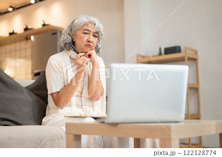 A perplexed Asian senior woman faces a problem while using her laptop, sitting in the living room. 122128774