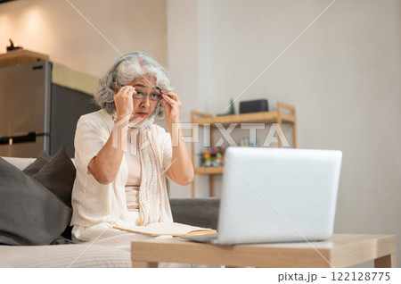 A senior woman gently wears her eyeglasses to read an article on the internet through her laptop. 122128775
