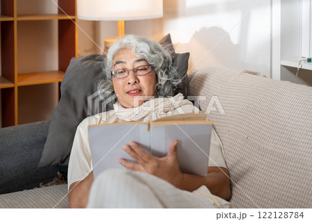 A relaxed Asian senior woman lies down on a sofa in the living room, reading a book at nighttime. 122128784