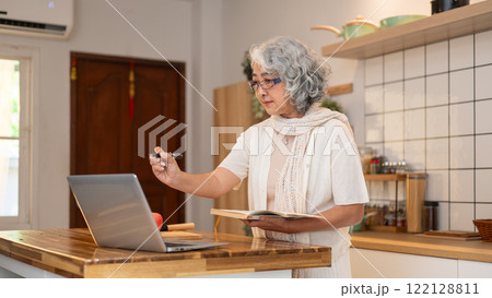 A positive retired elderly Asian woman learns a new recipe from the internet in the kitchen. A positive retired elderly Asian woman learns a new recipe from the internet in the kitchen. 122128811