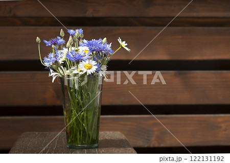 A bouquet of daisies and cornflowers in a glass vase on a wooden table against the background of a wooden wall. Place for an inscription. Selective focus. Copyspace. 122131192