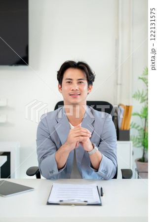 A successful Asian businessman sits at his desk in a modern office, smiling at the camera. 122131575