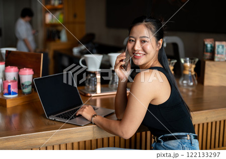 Attractive Asian woman sits at a counter bar in a coffee shop talking on the phone, working remotely Attractive Asian woman sits at a counter bar in a coffee shop talking on the phone, working remotely 122133297