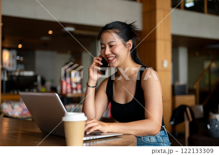 A confident Asian woman is talking on the phone while working on her laptop, working remotely. A confident Asian woman is talking on the phone while working on her laptop, working remotely. 122133309