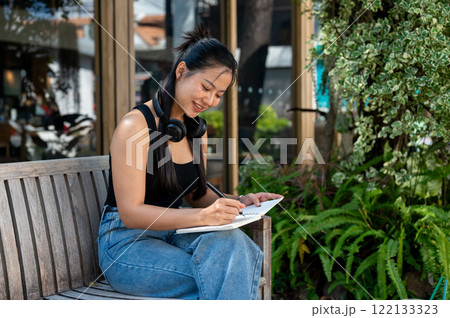 A beautiful, contemplating Asian woman writing her diary while relaxing on an outdoor bench. 122133323