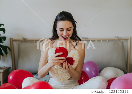 Beautiful woman sitting on bed in the middle of baloons, opening gift. Concept of Valentine's Day, love and romance. 122135331