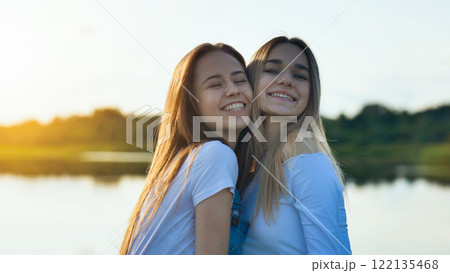 Two school girlfriends are embracing and smiling by the lake at sunset, enjoying their time together Two school girlfriends are embracing and smiling by the lake at sunset, enjoying their time together 122135468