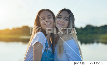 Teenage friends hugging, laughing near lakeside during golden sunset light, sharing warm friendship moment 122135480