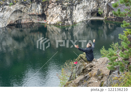 Young man sitting on a rock with raised arms, enjoying the view of a flooded marble quarry Young man sitting on a rock with raised arms, enjoying the view of a flooded marble quarry 122136210